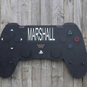 May include: A black wooden PlayStation controller-shaped wall art piece with the name "MARSHALL" in white. It features button symbols and a PlayStation logo. The art is mounted on a weathered wooden fence.