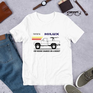 May include: White t-shirt with a black and white illustration of a Toyota Hilux pickup truck with a machine gun mounted on the bed. The text "TOYOTA HILUX" is above the truck and "FOR REGIME CHANGES ON A BUDGET" is below the truck.