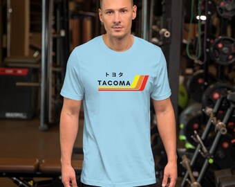 May include: Light blue t-shirt with a retro-style graphic design featuring the word "TACOMA" in black text with a yellow, orange, and red stripe design. The Japanese word for "Tacoma" is written in black above the word "TACOMA".