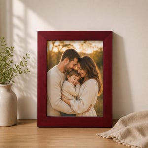 Peut inclure: Un cadre photo rectangulaire au fini rouge profond et lisse contenant une photographie de deux adultes et d'un jeune enfant s'enlaçant en extérieur sous une lumière dorée. Le cadre présente une bordure large et plate sur une surface en bois.