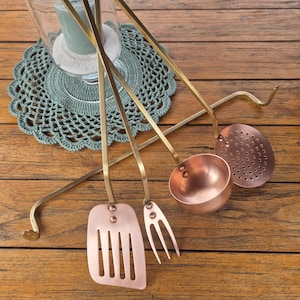May include: A set of copper and brass kitchen utensils, including a spatula, fork, ladle, and slotted spoon. The utensils have long brass handles and copper heads. The items are arranged on a wooden surface.