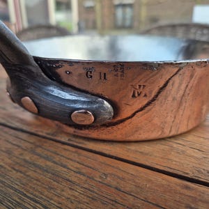 Peut inclure: Une casserole en cuivre avec une poignée en fer noir. La casserole porte les lettres G II et un N M stylisé estampés sur le fond.