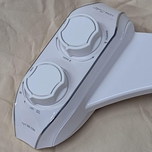 May include: Close-up of a white bidet toilet seat with control knobs. The knobs are labeled "Wash", "Max", "Off", "Feminine", and "Luxe Bidet".