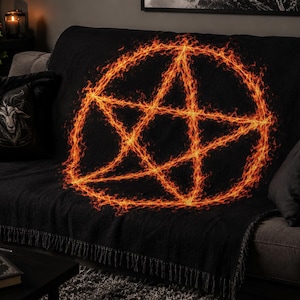 May include: Black throw blanket featuring a fiery orange pentagram design. The blanket is draped over a gray sofa, with a matching pillow featuring a goat head graphic. The fringe detail adds texture to the blanket.