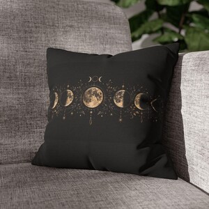 Puede incluir: Un cojín cuadrado negro con un gráfico horizontal central de las fases lunares en tonos dorados y beige. El diseño incluye ilustraciones lunares detalladas, delicados acentos de hojas botánicas, pequeñas estrellas y formas de cristal colgantes.