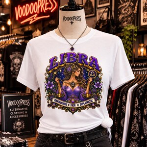 Könnte beinhalten: Ein weißes T-Shirt mit einer Grafik einer Frau, die eine Waage hält, als Symbol für das Sternzeichen Waage. Das Design enthält den violetten Schriftzug LIBRA und HARMONIOUS FAIR DIPLOMATIC, umrahmt von goldenen Verzierungen und lila Blumen.