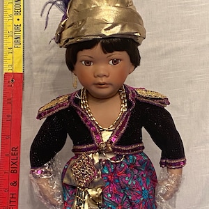 May include: A porcelain doll with dark brown hair and brown eyes, wearing a gold and purple outfit with a gold turban and a purple feather. The doll is wearing a long skirt with a floral pattern.
