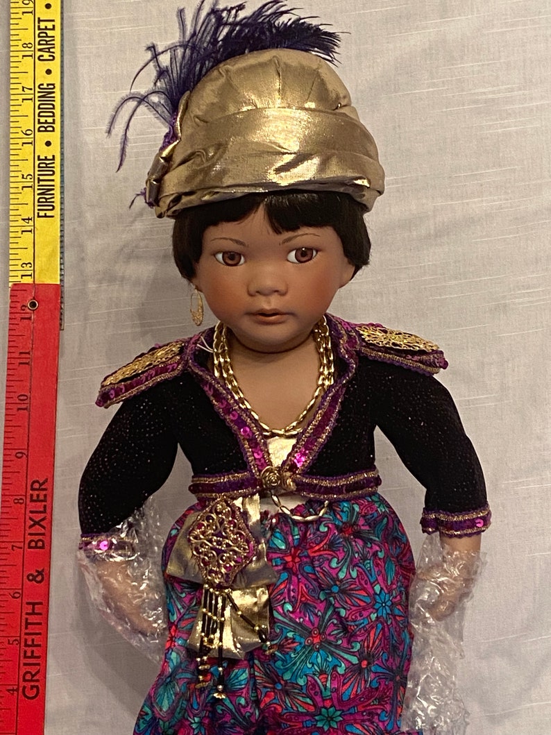 May include: A porcelain doll with dark brown hair and brown eyes, wearing a gold and purple outfit with a gold turban and a purple feather. The doll is wearing a long skirt with a floral pattern.