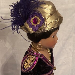 May include: A black doll with dark brown hair wearing a gold and purple outfit with a gold hat adorned with a purple feather and a jeweled embellishment.