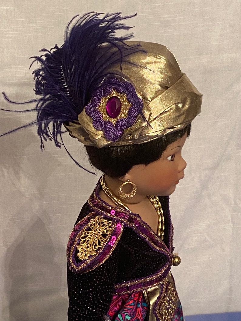 May include: A black doll with dark brown hair wearing a gold and purple outfit with a gold hat adorned with a purple feather and a jeweled embellishment.
