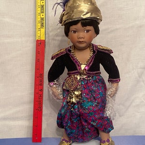 May include: A porcelain doll with dark brown skin, wearing a gold and purple outfit with a patterned skirt. The doll is wearing a gold turban with a purple feather and gold shoes.