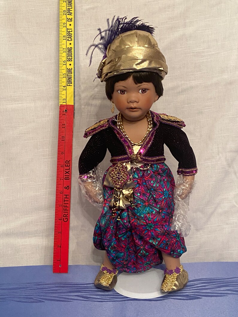 May include: A porcelain doll with dark brown skin, wearing a gold and purple outfit with a patterned skirt. The doll is wearing a gold turban with a purple feather and gold shoes.