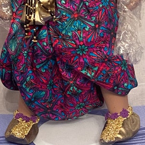 May include: A pair of gold shoes with purple trim and gold embellishments. The shoes are worn by a doll with a pink and blue patterned dress.