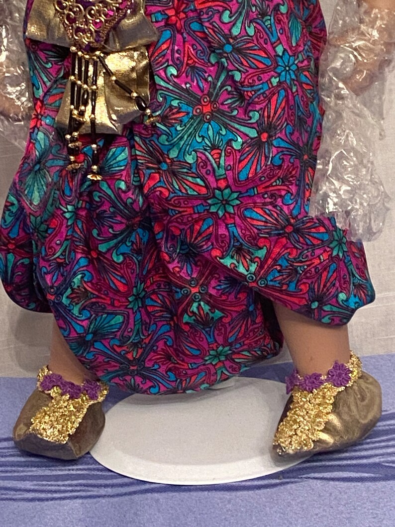 May include: A pair of gold shoes with purple trim and gold embellishments. The shoes are worn by a doll with a pink and blue patterned dress.