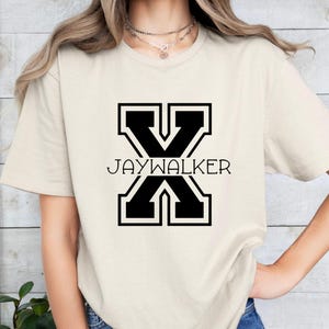May include: A beige t-shirt with a black graphic of a large letter X with the text "JAYWALKER" underneath.