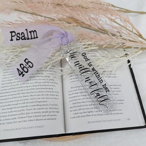 May include: A silver glitter bookmark with the text "God is within her, she will not fall" and the Bible verse "Psalm 46:5". The bookmark is decorated with a purple ribbon.