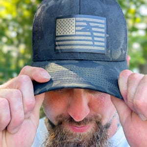 May include: Black camouflage baseball cap with an American flag patch. The flag features a tan and gold color scheme. The cap is being held up by a person.