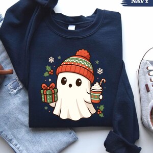 May include: Navy blue sweatshirt featuring a cartoon ghost wearing a red and green knit hat. The ghost holds a gift and a coffee cup with a candy cane. The design includes snowflakes and holly, suggesting a holiday theme.