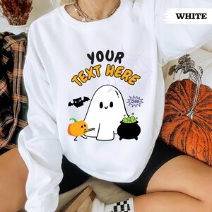 Puede incluir: Sudadera blanca con temática de Halloween con un fantasma, calabaza, murciélago y caldero. El texto "YOUR TEXT HERE" está en amarillo y negro. También hay un diseño de telaraña.