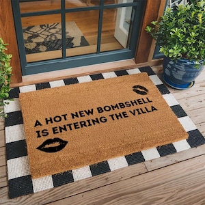 May include: A rectangular tan coir doormat featuring the black text A HOT NEW BOMBSHELL IS ENTERING THE VILLA with a black lip print icon positioned above the text on the right side and a larger black lip print icon below the text on the left side.