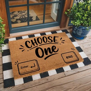 May include: A rectangular tan coir doormat featuring the black text "CHOOSE ONE" centered above two black graphic icons resembling computer keyboard keys, one labeled "Enter" with an arrow symbol and the other labeled "esc".