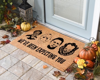 May include: A brown coir doormat with the text "We've Been Expecting You" and four black and white horror movie character faces. The faces are of Michael Myers, Jason Voorhees, Freddy Krueger, and Pennywise.