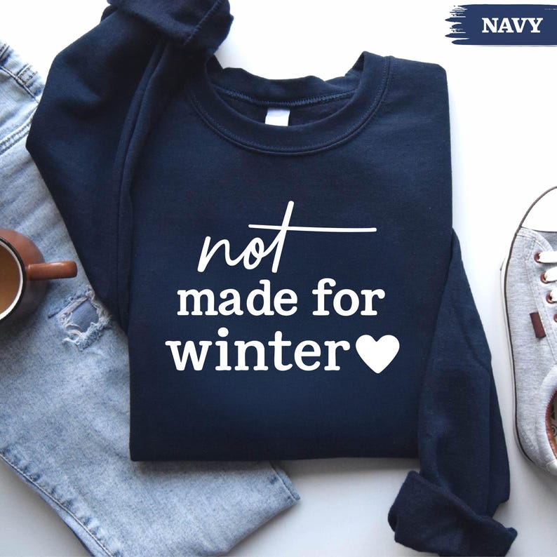 Puede incluir: Sudadera azul marino con el texto blanco "not made for winter" y un coraz&oacute;n blanco. La sudadera se muestra sobre una superficie de mezclilla azul claro, con una taza de caf&eacute; y zapatillas grises.