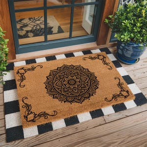 May include: A rectangular doormat with a brown coir surface features a black mandala design in the center and decorative corner accents. It sits atop a black and white checkered mat, near a door and potted plant.