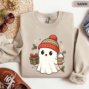 Puede incluir: Sudadera color arena con un fantasma de dibujos animados que lleva un gorro de punto rojo y verde. El fantasma sostiene un regalo envuelto y una taza de café con un bastón de caramelo. Acentos festivos de acebo y copos de nieve completan el diseño.