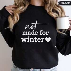Puede incluir: Sudadera negra con el texto blanco "not made for winter" y un coraz&oacute;n blanco. La palabra "black" est&aacute; en la esquina superior derecha. Una taza blanca se sostiene en la mano derecha.
