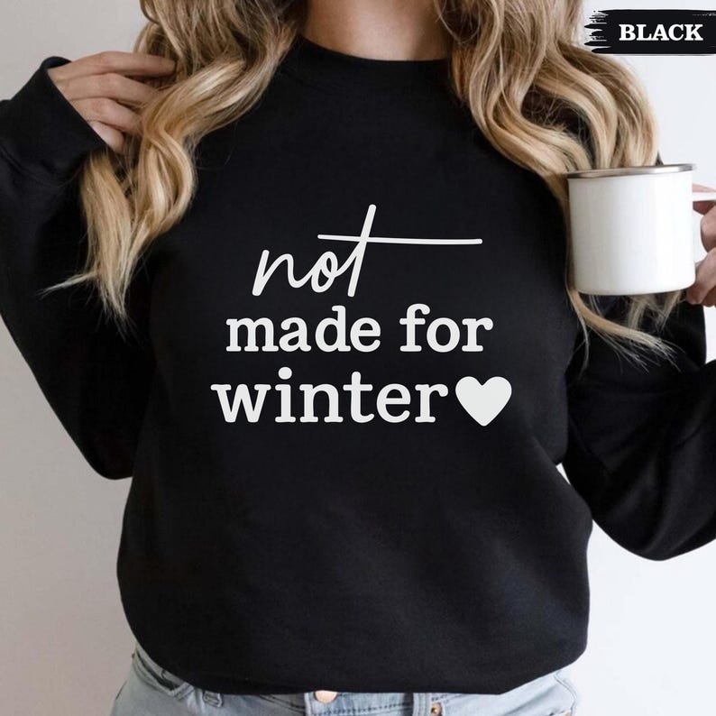 Puede incluir: Sudadera negra con el texto blanco "not made for winter" y un coraz&oacute;n blanco. La palabra "black" est&aacute; en la esquina superior derecha. Una taza blanca se sostiene en la mano derecha.