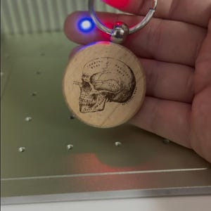 May include: A wooden keychain with a laser-engraved skull design. The keychain is attached to a silver metal ring.
