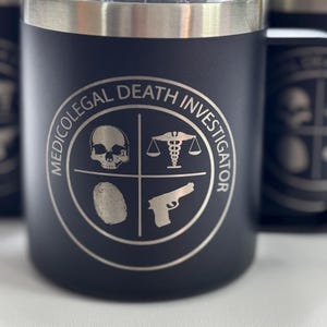 May include: Black stainless steel travel mug with a silver logo that says "Medico-Legal Death Investigator" with four icons: a skull, a scale with a caduceus, a fingerprint, and a gun.