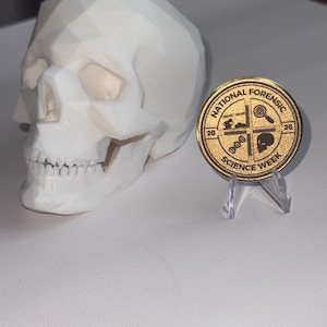 May include: A white low-poly geometric skull sculpture positioned next to a circular gold-colored commemorative coin on a clear plastic stand. The coin features the text "NATIONAL FORENSIC SCIENCE WEEK" with icons of a magnifying glass, DNA, and a skull.