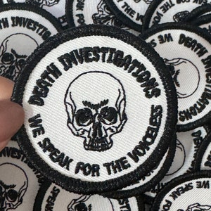 May include: Round white and black patches with a skull graphic. The patches have the text "DEATH INVESTIGATIONS" and "WE SPEAK FOR THE VOICELESS" in a circular design. The patches are embroidered.