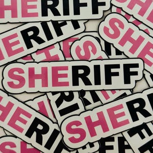 May include: A collection of white stickers with the word "SHERIFF" printed in black letters with a pink outline. The word "SHERIFF" is also printed in pink letters.