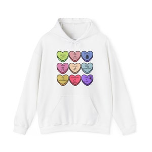 Könnte beinhalten: Weißer Hoodie mit einem Motiv aus neun bunten Bonbonherzen, jedes mit einer anderen Botschaft. Die Botschaften lauten: "Selbstpflege ist so schick", "Du bist ein guter Mensch", "Selbstmedikation ist keine Therapie", "Lass uns nicht Freunde sein", "Iss den gelben Schnee nicht", "Wir haben alle unsere eigenen Probleme", "Nicht schlecht für einen Menschen", "Eigentlich bist du derjenige, der verrückt ist", und "Es ist okay, manchmal traurig zu sein".