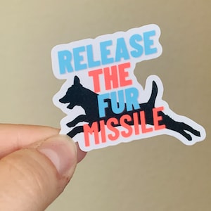 May include: A sticker featuring a black silhouette of a dog in mid-run. The sticker has the text "RELEASE THE FUR MISSILE" in blue and red, with a white border. The sticker is held by a hand.