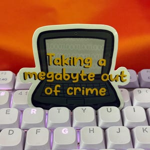 May include: A sticker of a laptop with the text "Taking a megabyte out of crime" on a white keyboard with purple backlighting.