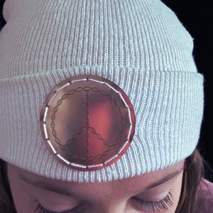 May include: White knit beanie with a peace sign pin featuring a DNA double helix design. The pin is made of metal and has a copper finish.