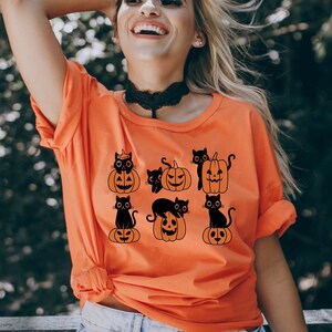 May include: Orange t-shirt with a black print of cats sitting on pumpkins. The cats are wearing witch hats and have spooky expressions.