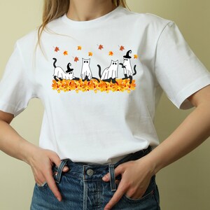 May include: White t-shirt with a graphic of four cats dressed as ghosts and witches in a pile of orange and yellow leaves.