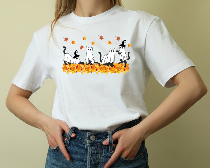 May include: White t-shirt with a graphic of four cats dressed as ghosts and witches in a pile of orange and yellow leaves.