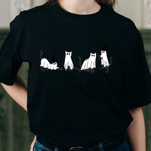May include: Black t-shirt with a graphic of five white cats dressed as ghosts. One cat is wearing a witch's hat.