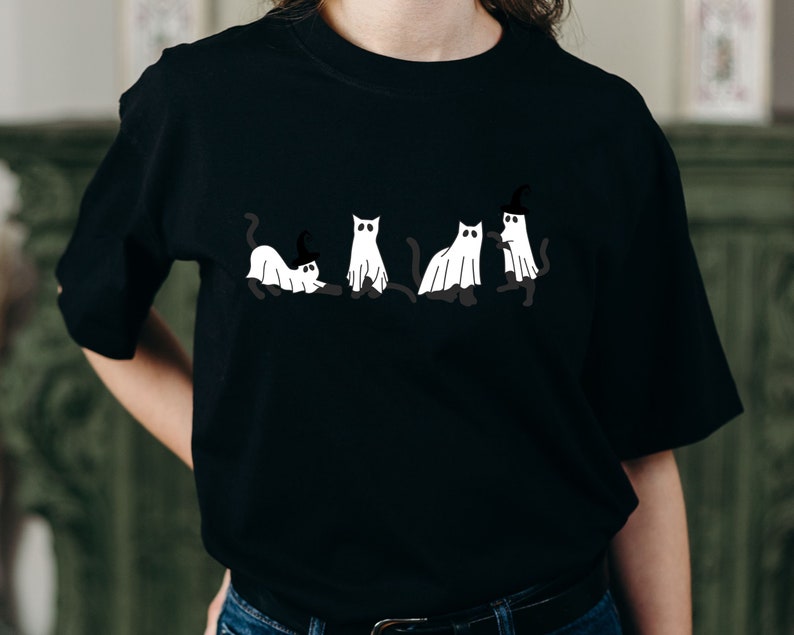 May include: Black t-shirt with a graphic of five white cats dressed as ghosts. One cat is wearing a witch's hat.