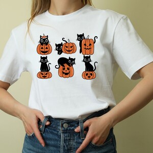 May include: White t-shirt with a black cat and pumpkin design. The design features multiple black cats sitting on top of orange pumpkins with jack-o-lantern faces.