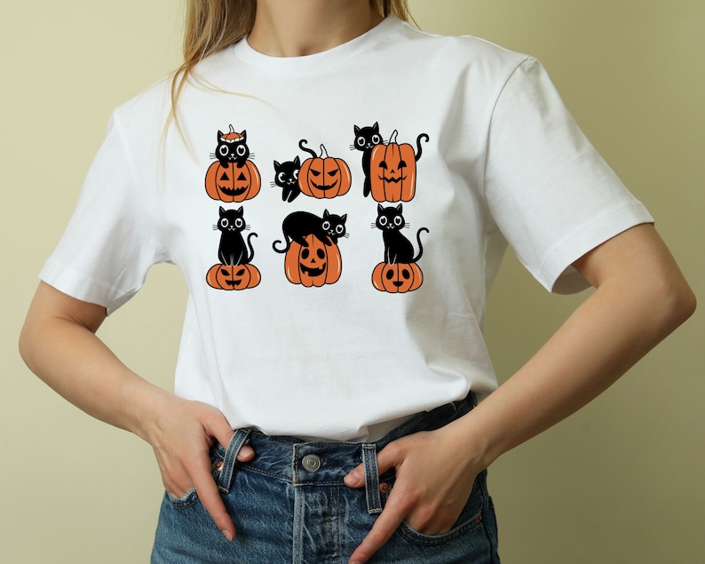 May include: White t-shirt with a black cat and pumpkin design. The design features multiple black cats sitting on top of orange pumpkins with jack-o-lantern faces.