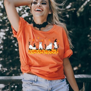 May include: Orange t-shirt with a graphic of four white cats dressed as ghosts and witches on a bed of orange and yellow leaves.