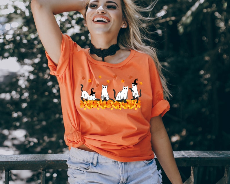 May include: Orange t-shirt with a graphic of four white cats dressed as ghosts and witches on a bed of orange and yellow leaves.