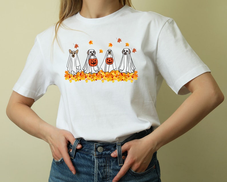May include: White t-shirt with a graphic of four dogs dressed as ghosts for Halloween. The dogs are walking in a line and are surrounded by orange and yellow fall leaves.